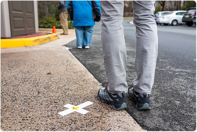 People standing waiting in line far apart to maintain social distance distancing during COVID-19 coronavirus outbreak with mark cross signs on sidewalk pavement by grocery food store. Image Credit: Kristi Blokhin