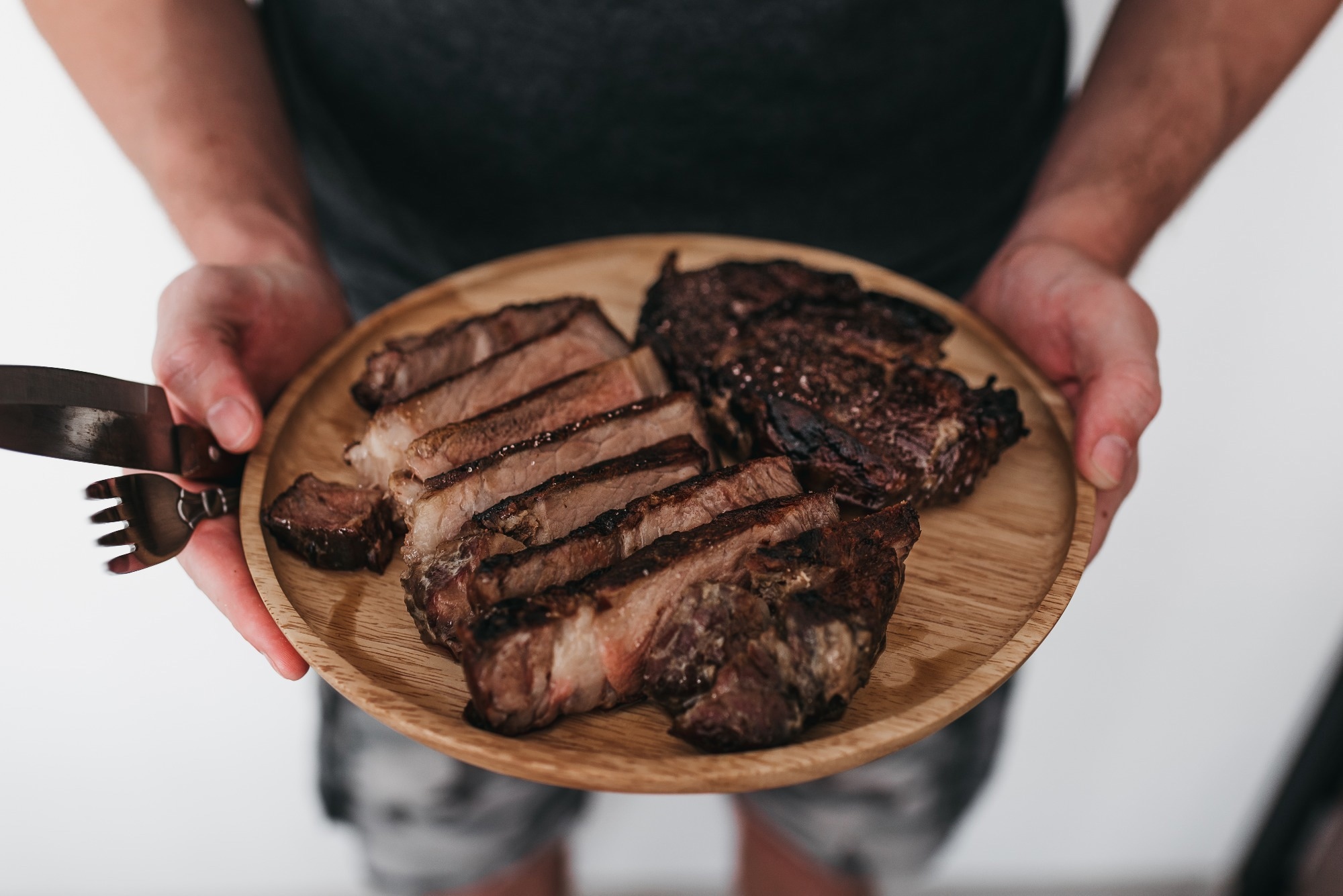 A man holding a wooden plate that has rib eye steak on it.