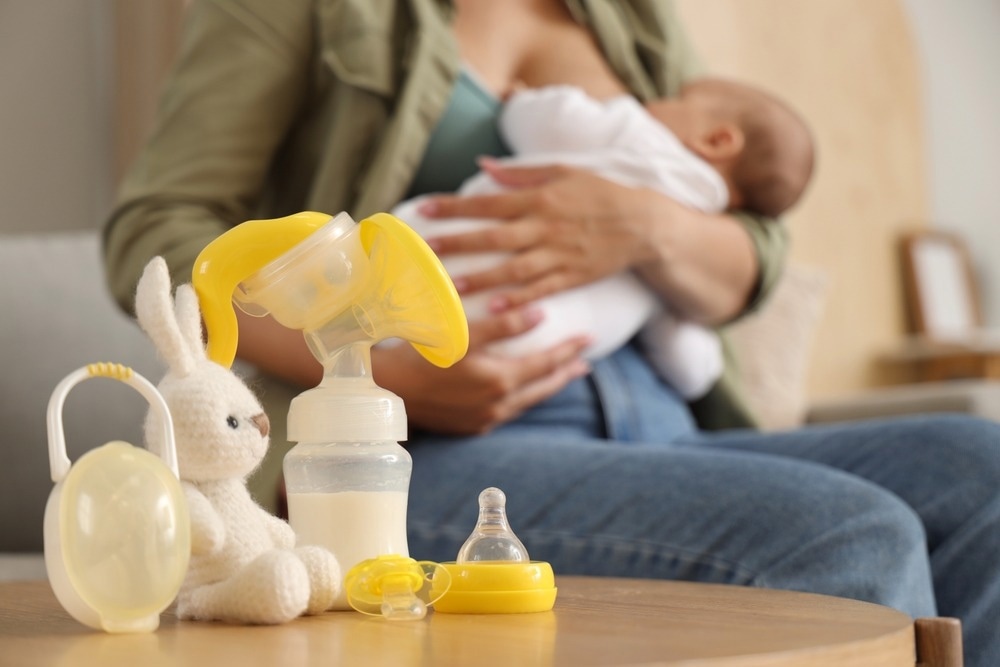 A breast pump, baby bottle, and pacifier are arranged on a wooden table next to a stuffed bunny, while a mother breastfeeds her baby in the blurred background.