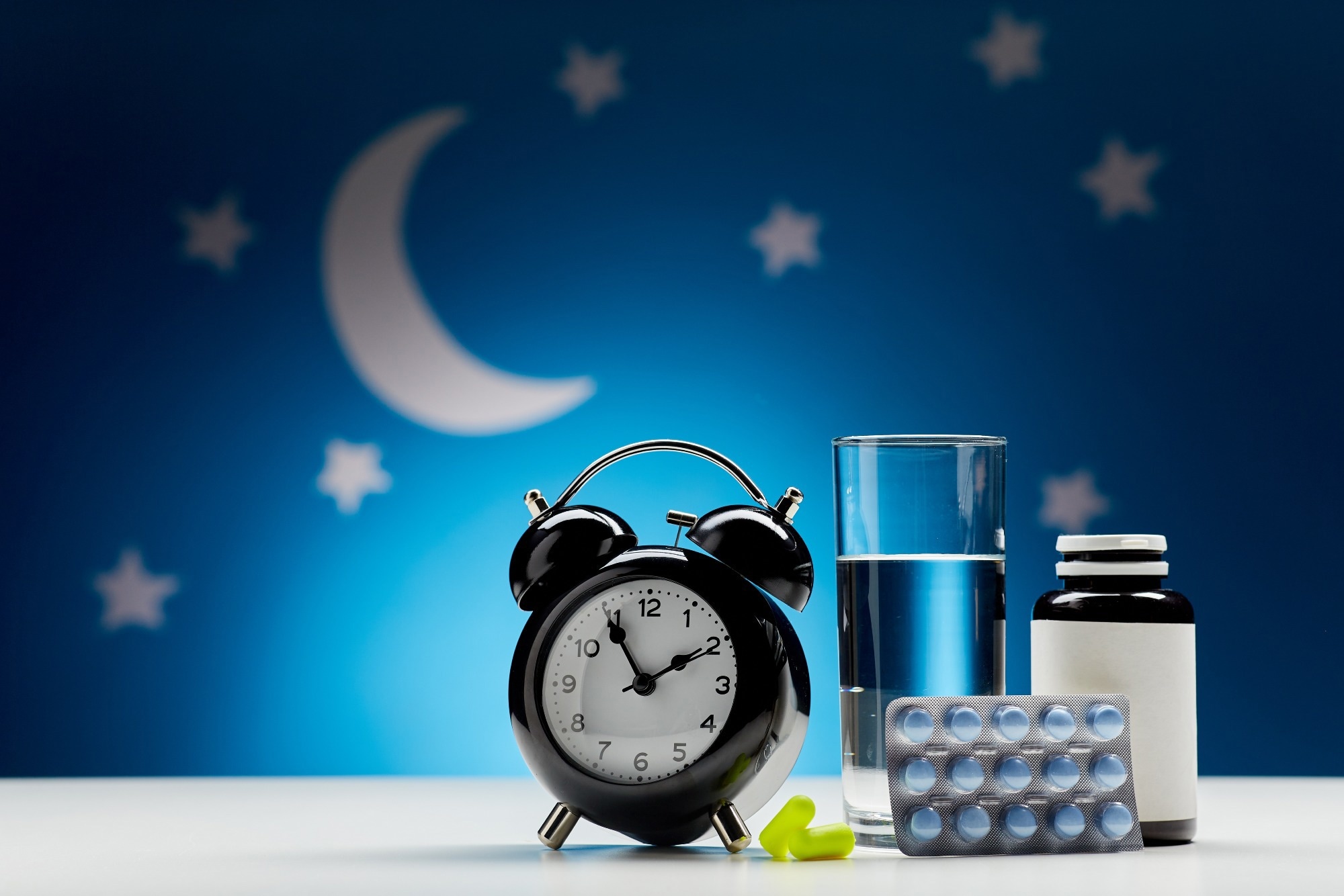 close up of alarm clock, glass of water, earplugs and sleeping pills over moon and night stars on blue background