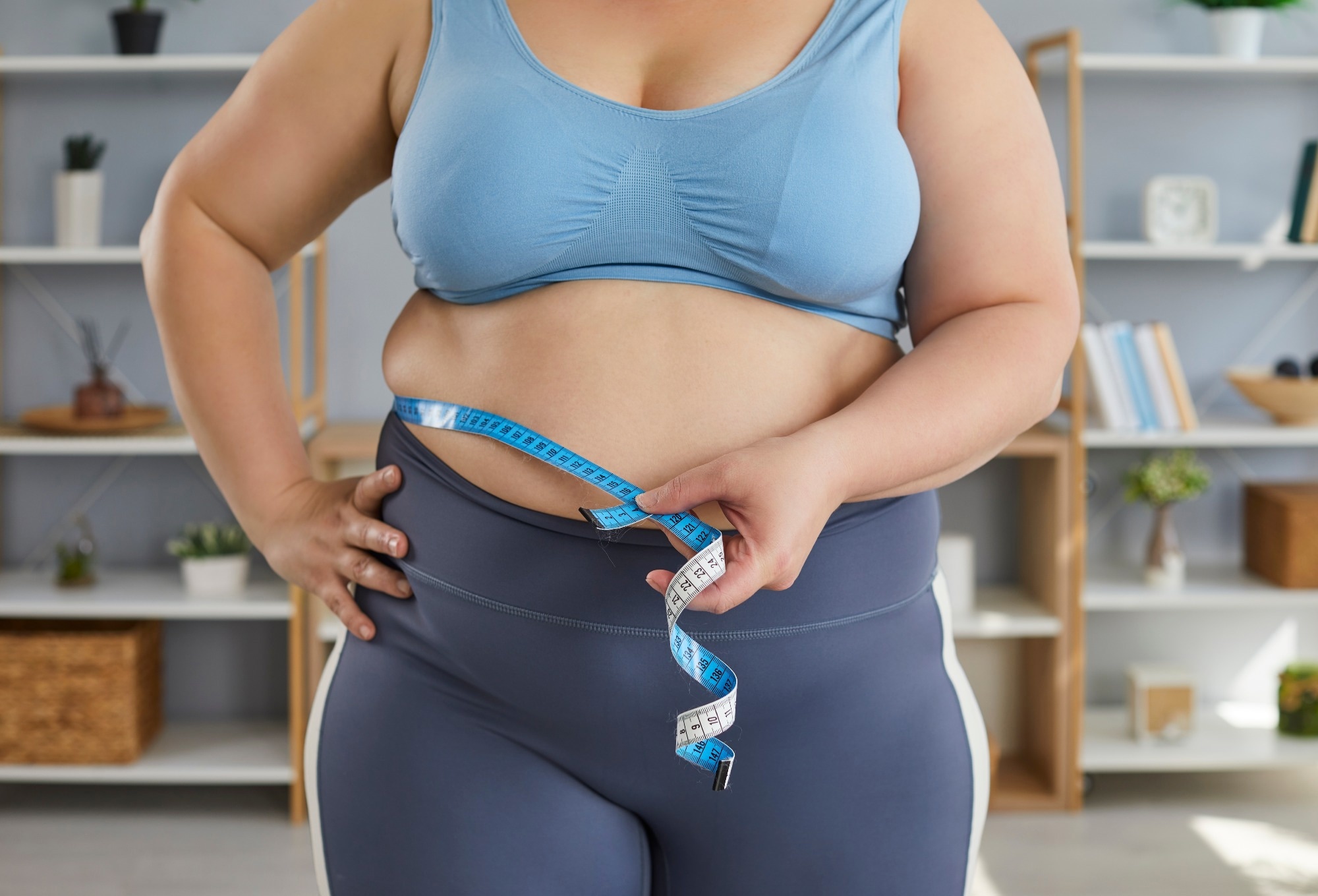 Close up cropped photo of a overweight woman wearing sportswear measure the waist with measuring tape standing in the living room at home.