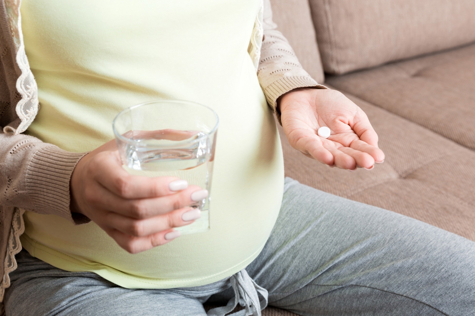 Close up of pregnant woman with tablet medicine in ones hand and a glass of water in the other hand. Sat on sofa at home.